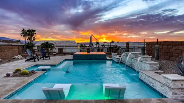 A swimming pool with a waterfall feature, surrounded by lounge chairs and palm trees, under a sunset sky.