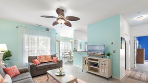 Een woonkamer met een bank, salontafel en televisie. Er is een plafondventilator, een keuken met een eettafel, en uitzicht op het strand.