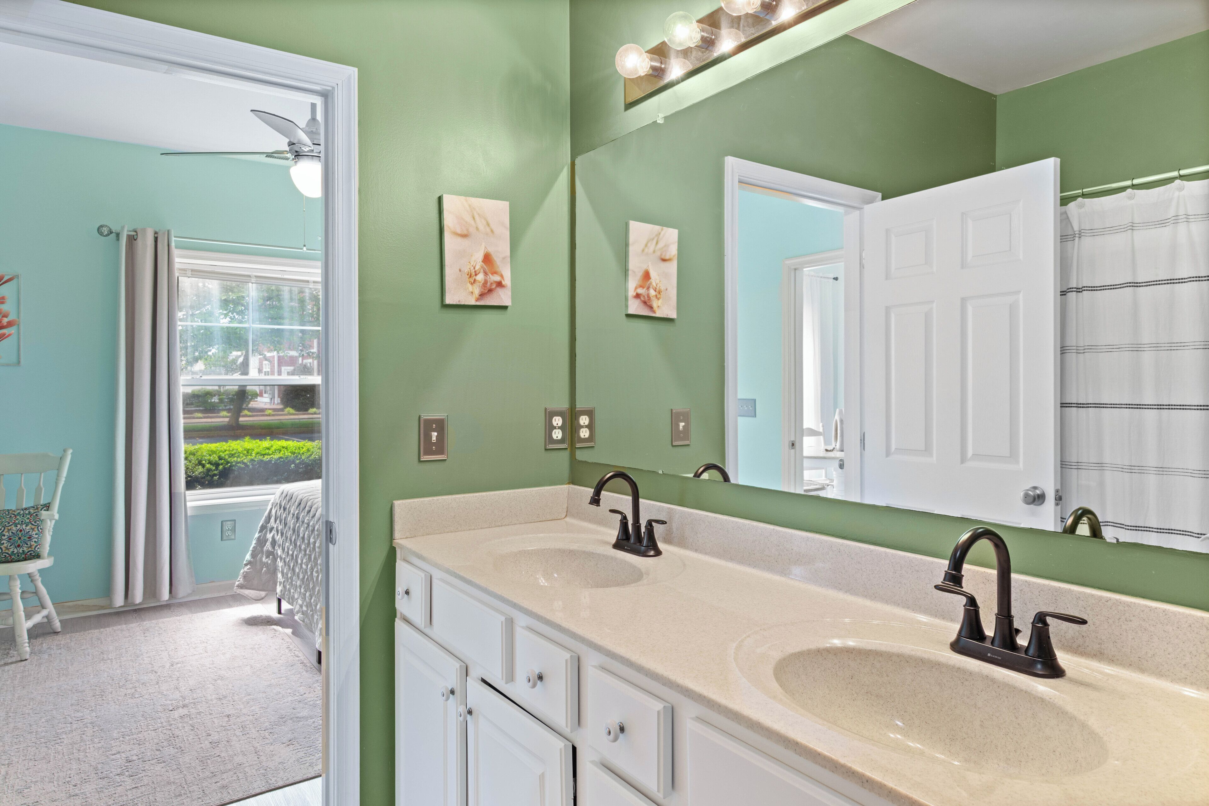A bathroom with two sinks, a large mirror, and a ceiling fan. There is a window with curtains and a view of greenery outside.