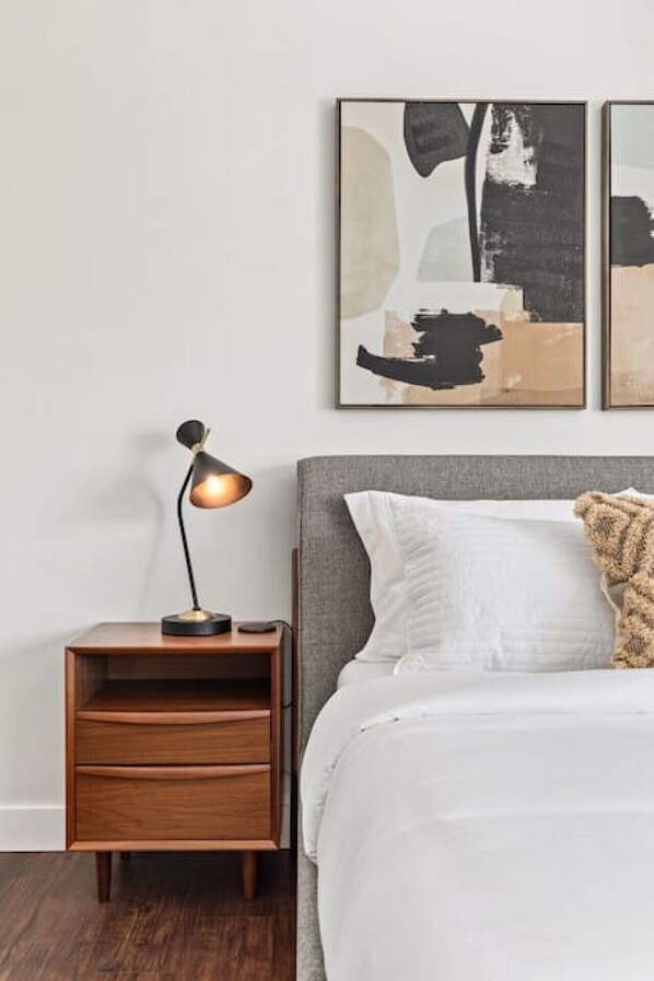 Une chambre avec une table de chevet en bois, une lampe de bureau, un lit avec une literie blanche et des œuvres d’art abstraites accrochées au mur.