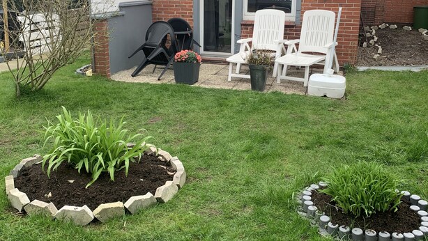 Un patio trasero con una casa de ladrillo, dos sillas blancas, una parrilla y dos bancales elevados con plantas.