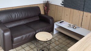 Una sala de estar moderna con un sofá de cuero marrón, una mesa de centro redonda de madera, un televisor de pantalla plana montado en una pared con diseño de ladrillo y una mesita auxiliar con objetos decorativos.
