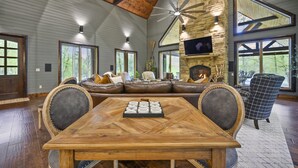 Un vaste salon doté d’une grande table en bois, de sièges confortables, d’une cheminée en pierre et de grandes fenêtres offrant une vue sur l’extérieur.
