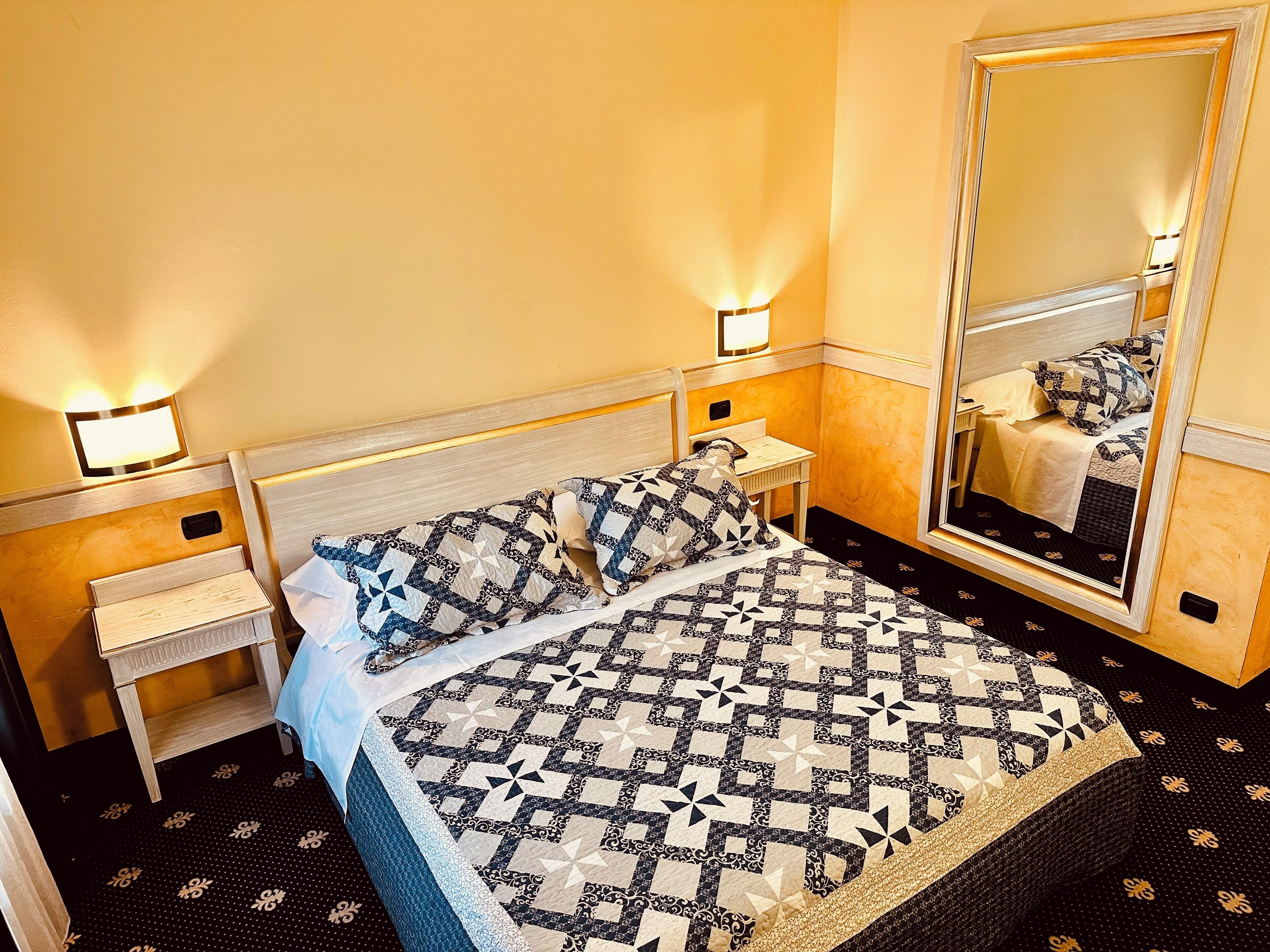 A hotel room with a bed, bedside tables, a mirror, and a wall-mounted light fixture.