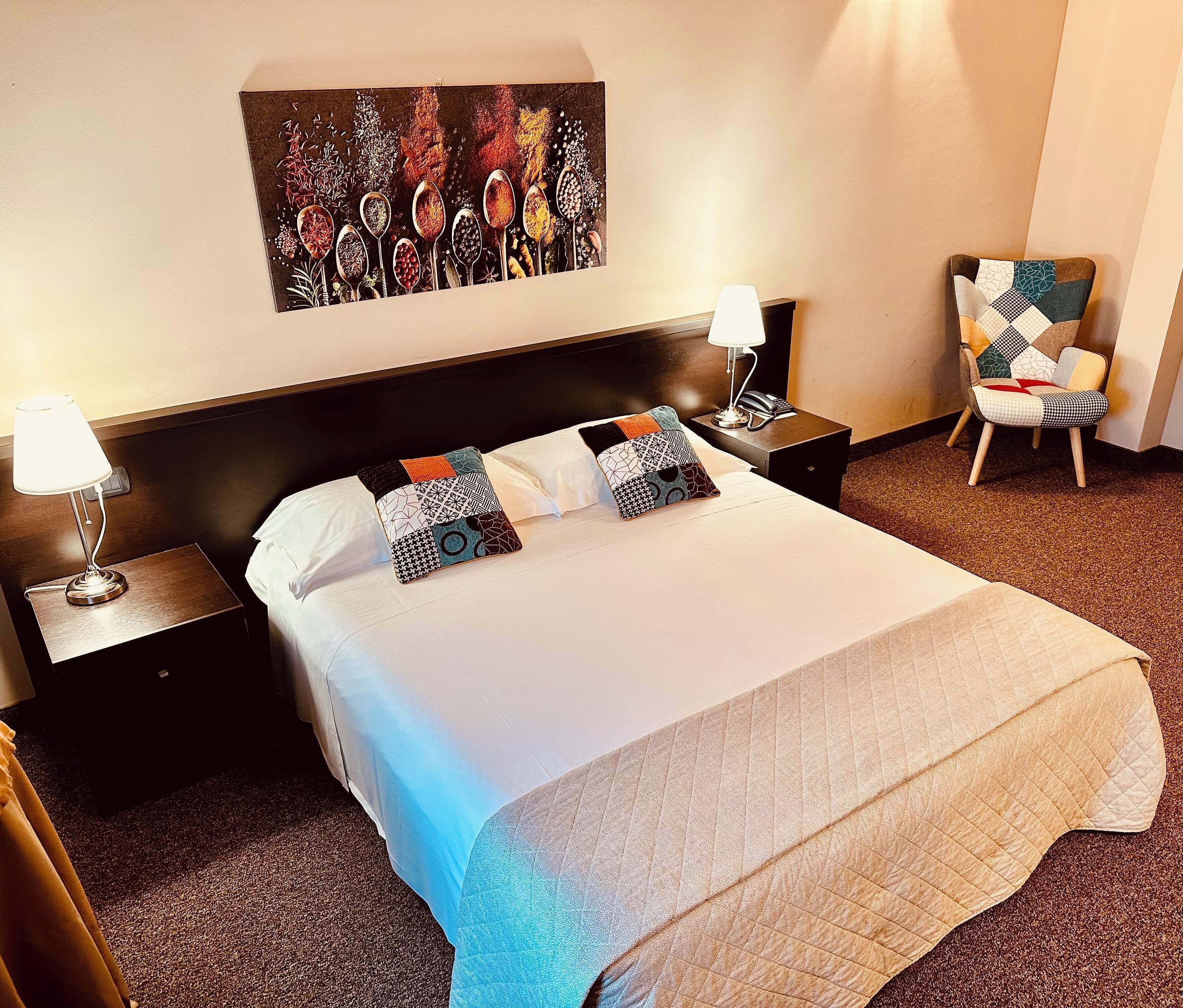 Una habitación de hotel con una cama grande, dos mesitas de noche con lámparas, una silla y una obra de arte en la pared.