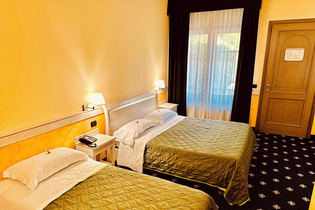 A hotel room with two beds, a nightstand with a phone, a window with sheer curtains, and a door with a sign.