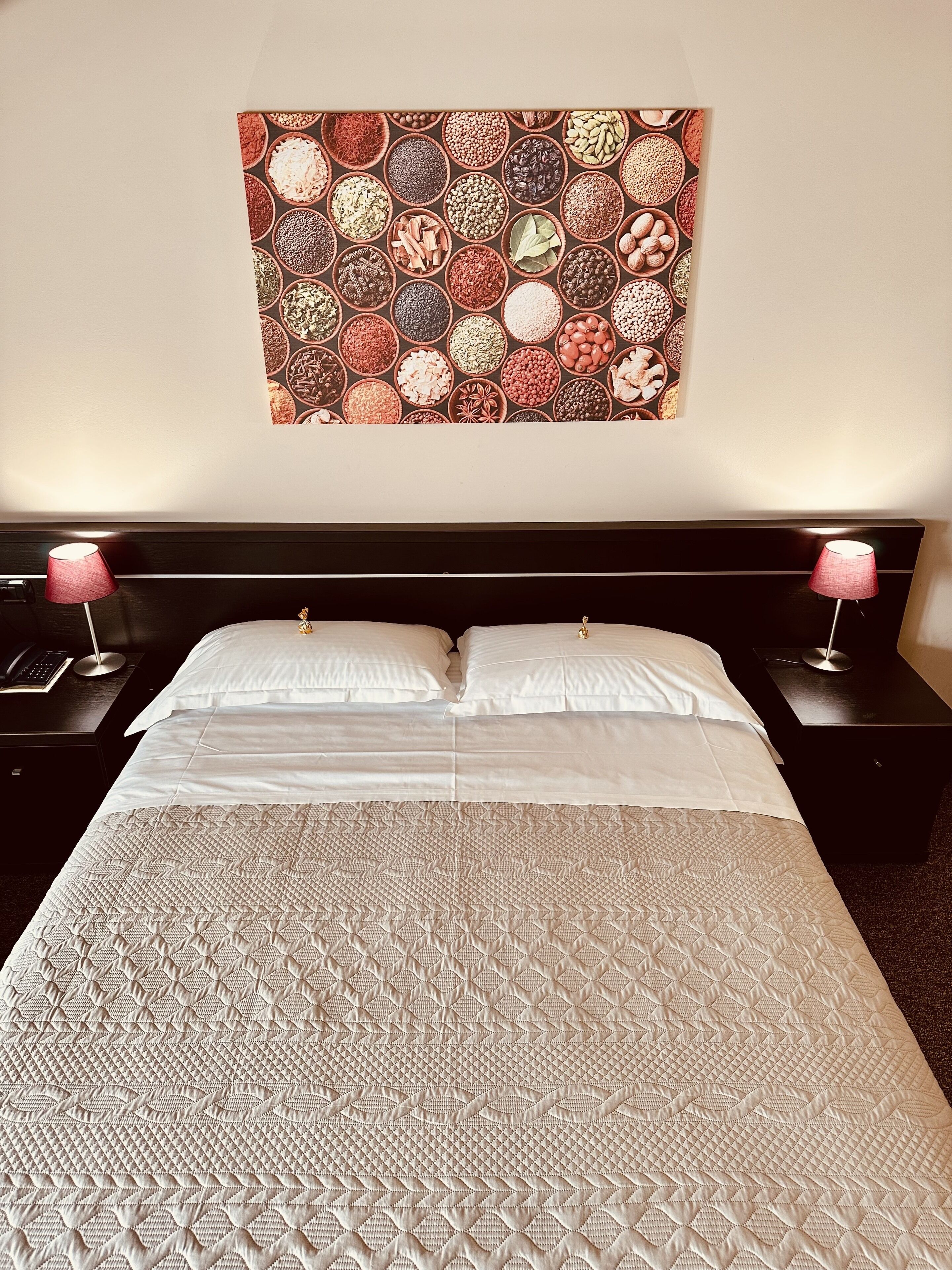 A neatly made bed with a patterned bedspread, two bedside lamps, and a wall tapestry of various spices.