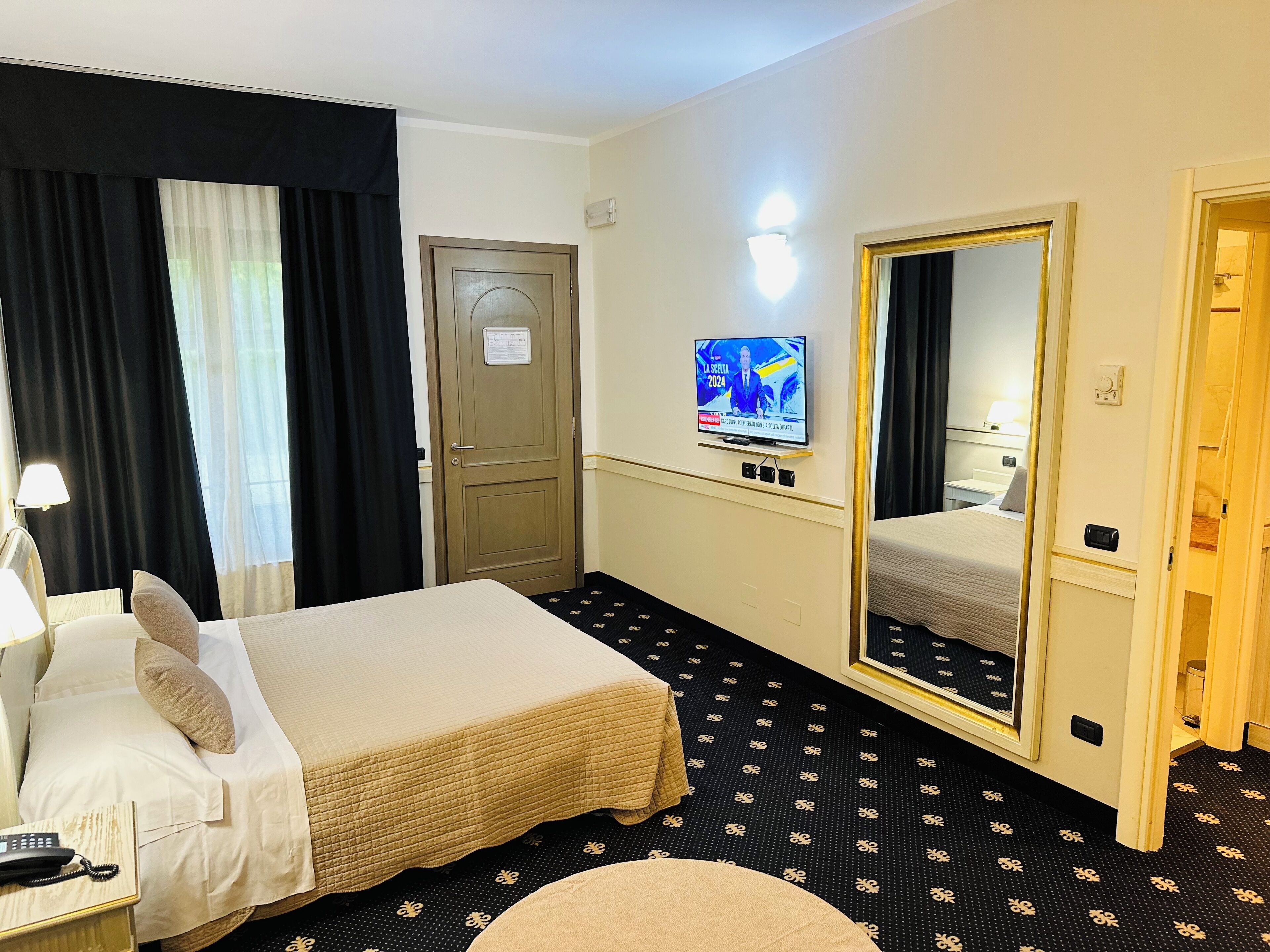 Una habitación de hotel con cama, escritorio con teléfono, televisor, espejo y puerta.