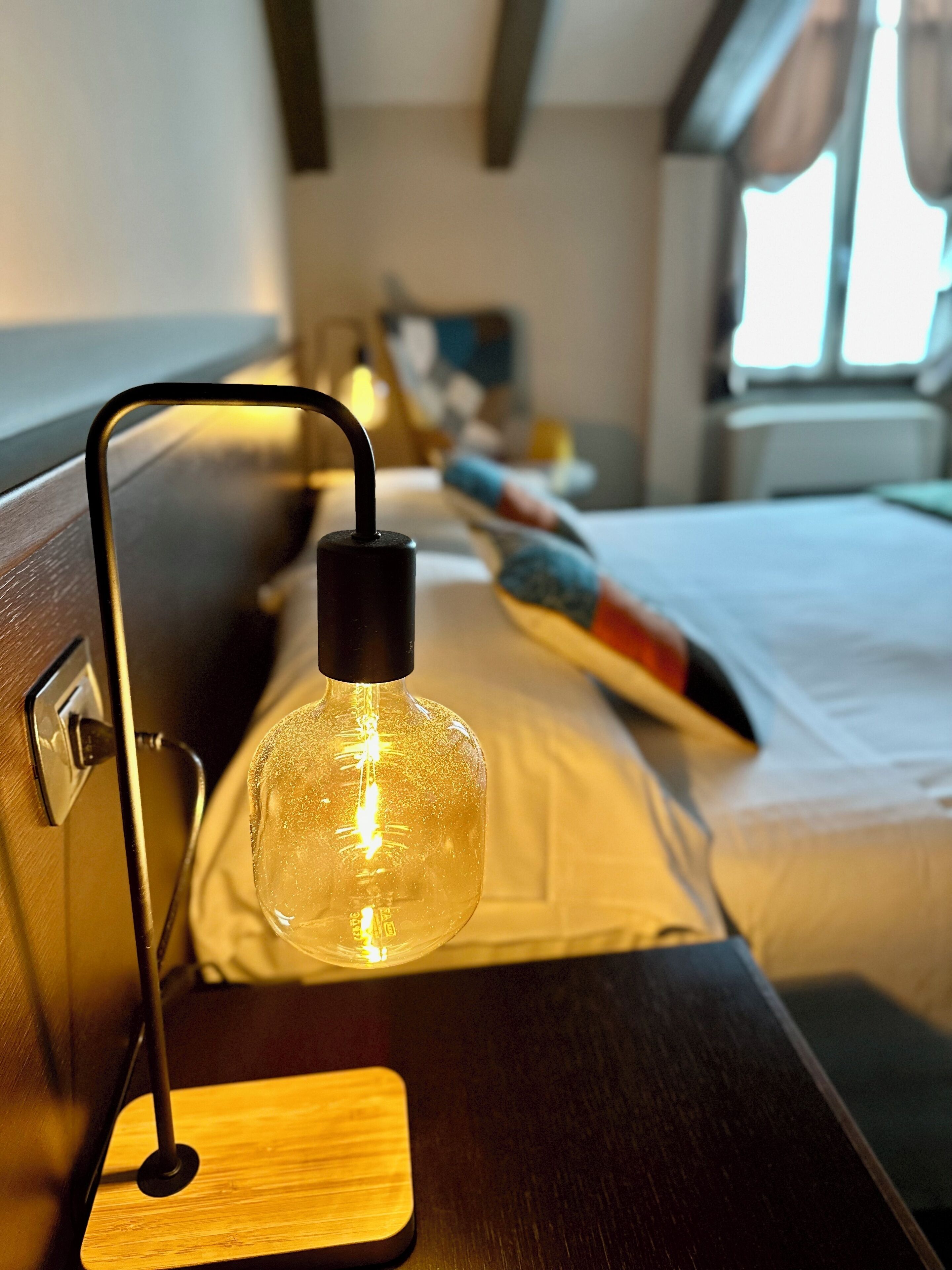 A bed with a wooden headboard, a bedside table with a lamp, and a window with curtains.