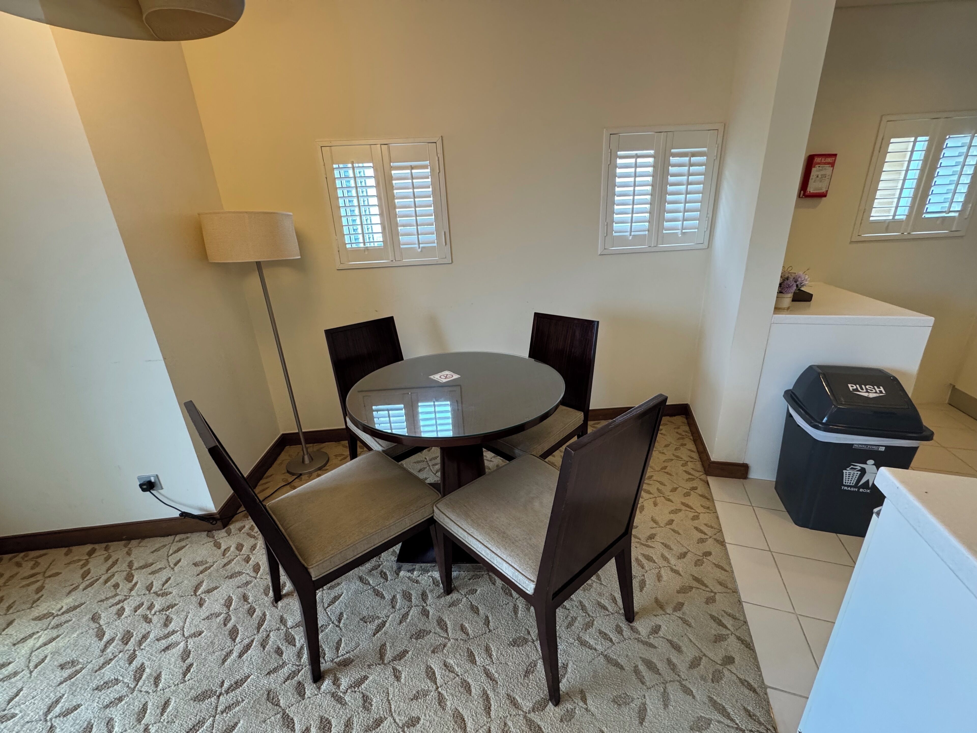 A dining area with a round table and four chairs, two windows with blinds, a lamp, and a trash bin.