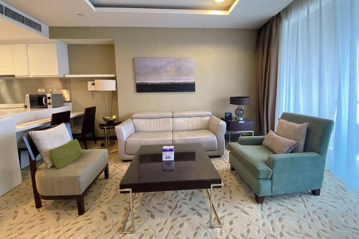 A modern hotel room with a living area featuring a sofa, two armchairs, a coffee table, a flat-screen TV, a dining area with a microwave, and a large window with curtains.