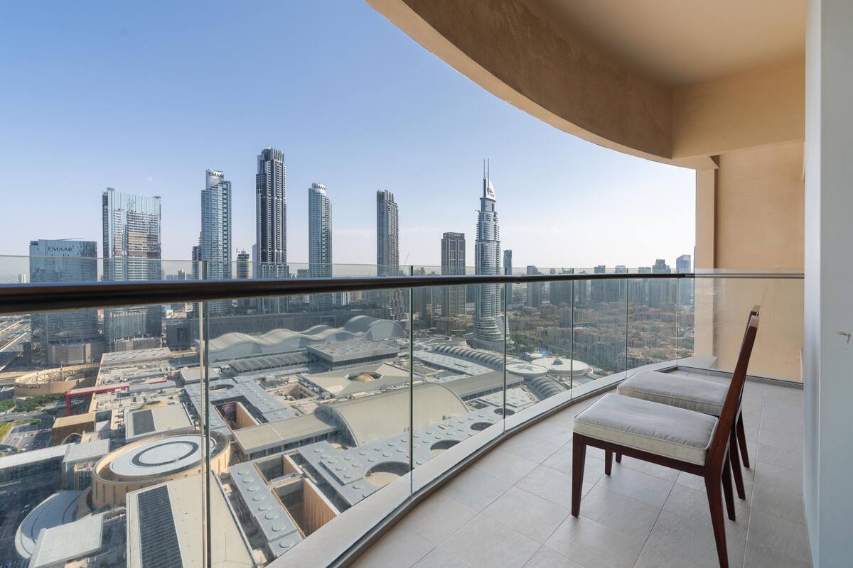 A high-rise balcony with a city view, featuring a glass railing, a cushioned chair, and a modern architectural setting.