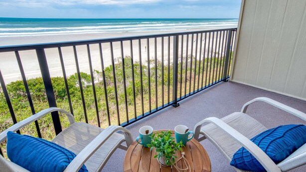 A balcony with a view of the beach, two chairs, a small table with a plant and two cups.