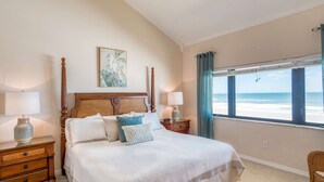 A bedroom with a large bed, two bedside tables, a nightstand, a chair, a window with a view of the beach, and a wall-mounted artwork.