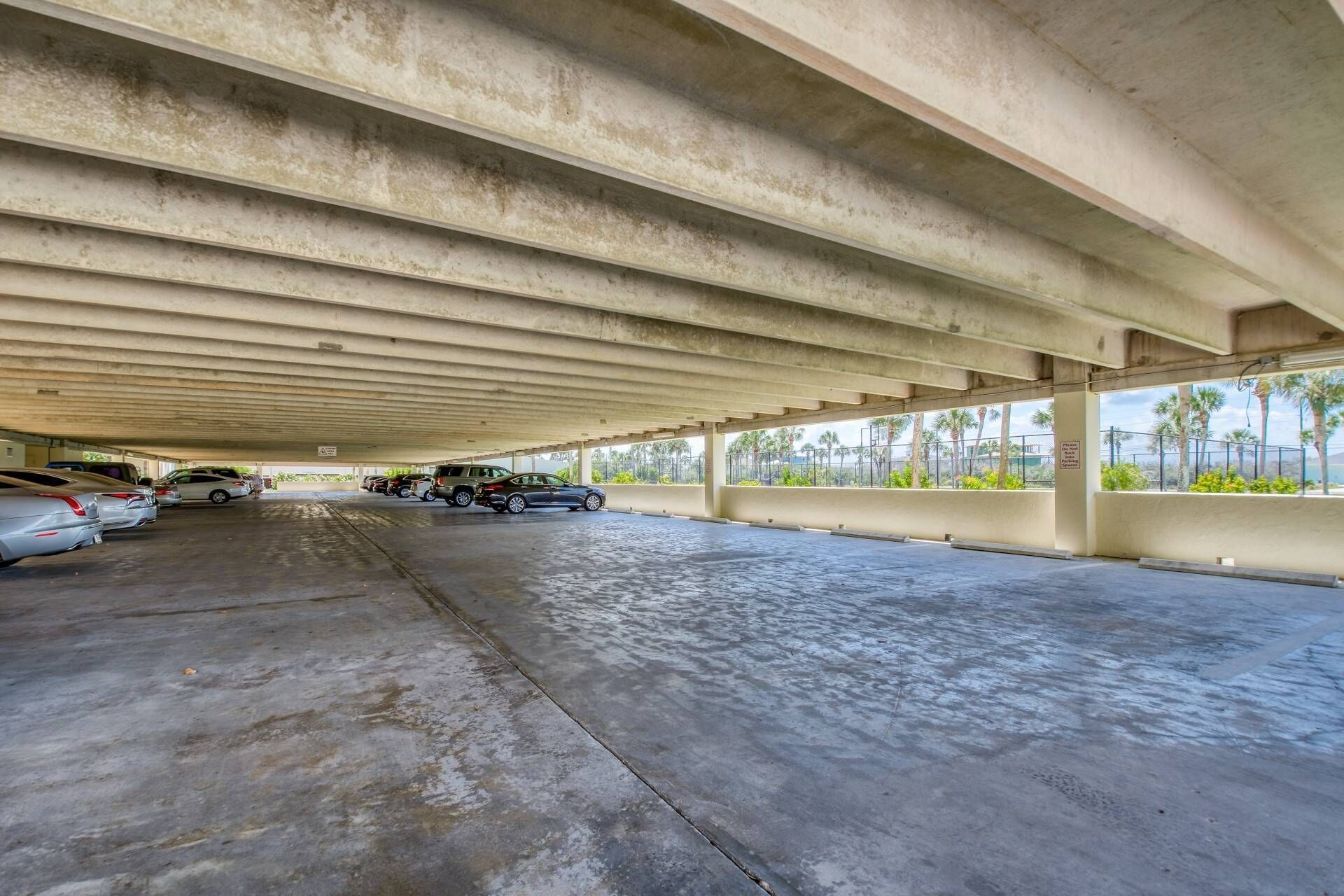 An underground parking garage with concrete ceilings and walls, featuring several parked cars and palm trees visible through the windows.