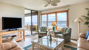 A living room with a TV, glass coffee table, floral armchairs, and a view of the ocean through large windows. - Sand Dollar #205 (St. Augustine)
