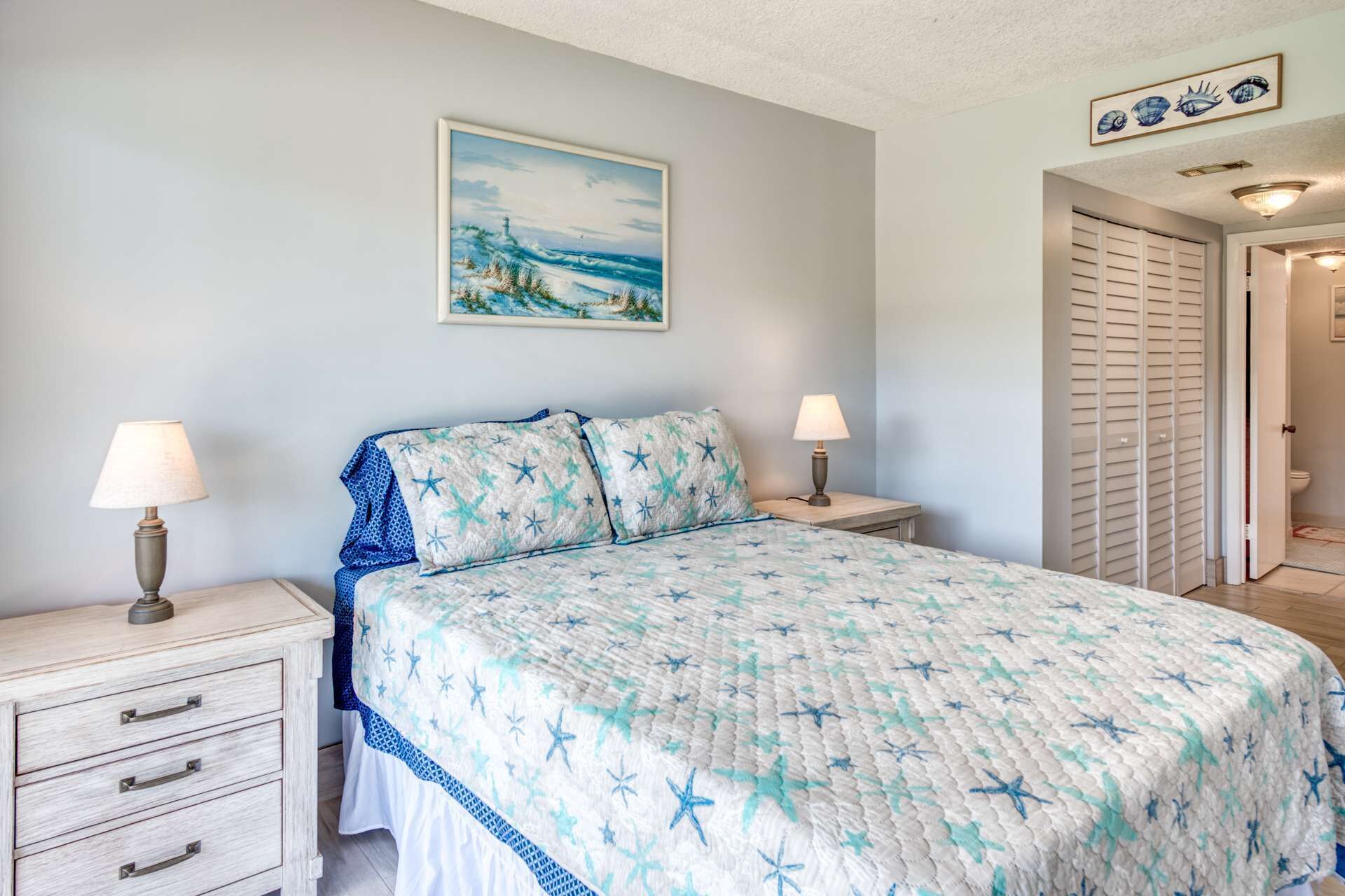 A bedroom with a bed, two bedside tables, a nightstand, a drawer unit, a painting on the wall, and a door leading to another room.