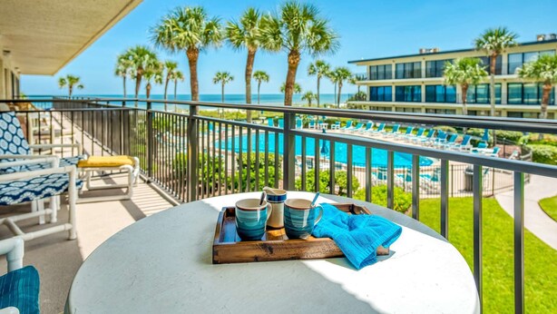 A balcony with a table set with blue and white mugs, a blue towel, and a wooden tray. In the background, there is a pool area with lounge chairs and palm trees, and a building with large windows. - Grandma's Rest East (St. Augustine)