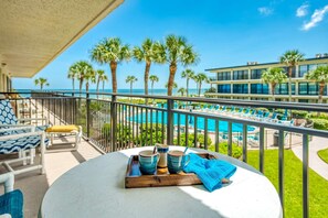 A balcony with a table set with blue and white mugs, a blue towel, and a wooden tray. In the background, there is a pool area with lounge chairs and palm trees, and a building with large windows. - Grandma's Rest East (St. Augustine)