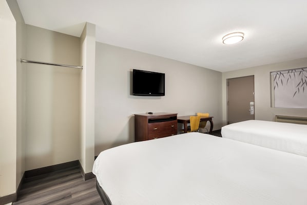 Habitación de hotel con dos camas, un televisor montado en la pared, un escritorio con una silla y una mesita con una lámpara.