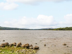 Ein ruhiger See mit steiniger Uferlinie und Hügeln im Hintergrund unter bewölktem Himmel.