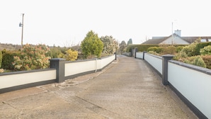 Un camino de concreto bordeado por paredes blancas y follaje, que conduce a una zona residencial con casas y árboles.