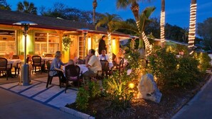 An outdoor dining area with tables and chairs, palm trees, and a building with warm lighting. - Beach Cottage with a Private Hot Tub (Longboat Key)