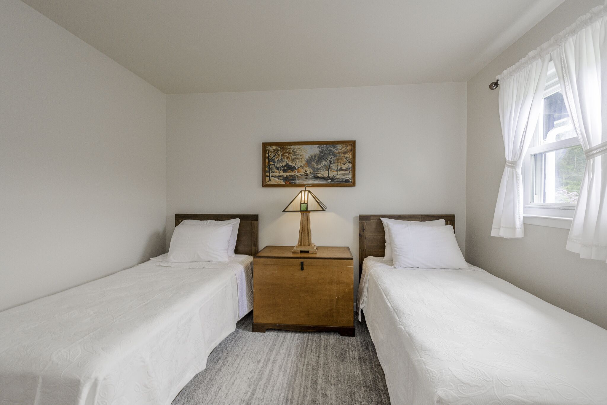 A hotel room with two single beds, a wooden nightstand with a lamp, a painting on the wall, and a window with curtains.