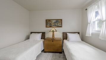 A hotel room with two single beds, a wooden nightstand with a lamp, a painting on the wall, and a window with curtains.