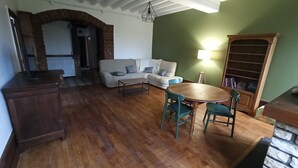 Ein Wohnzimmer mit Sofa, Couchtisch und Esstisch mit Stühlen. Es gibt ein Bücherregal und eine Lampe im Raum.