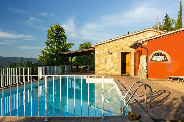 A swimming pool with a stone wall and a metal railing, a covered patio area, and a view of the mountains.