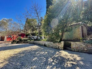Une cour pavée de pierres, bordée d’un mur de pierre, avec un bâtiment rouge et un arbre aux branches nues.