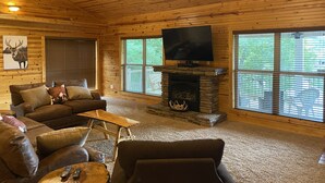 A cozy cabin interior with a stone fireplace, wooden walls, a ceiling fan, and a large window. - Large House with Front Row View of the Cliffs (Edgemont)