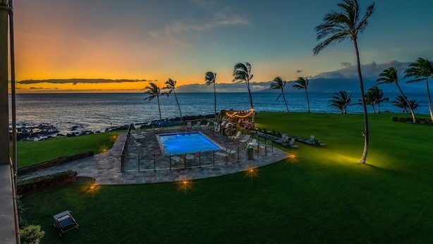 A pool area with a view of the ocean, surrounded by palm trees and a sunset. - 308 – Deluxe Oceanfront One Bedroom Unit with A/C! (Kihei)