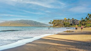 A beach with a hotel in the background, palm trees, and mountains in the distance. - 308 – Deluxe Oceanfront One Bedroom Unit with A/C! (Kihei)