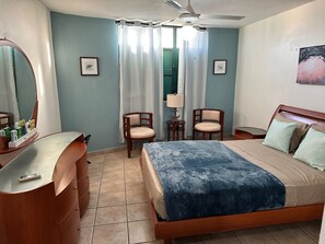 Ein Schlafzimmer mit einem Bett, zwei Stühlen, einem Schminktisch, einem Spiegel, einem Deckenventilator und einem Fenster mit Vorhängen.