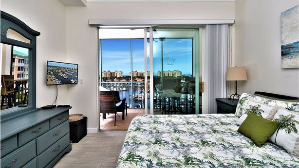 Habitación de hotel con una cama grande, un tocador, televisión, balcón con vistas a la ciudad y un cuadro en la pared.