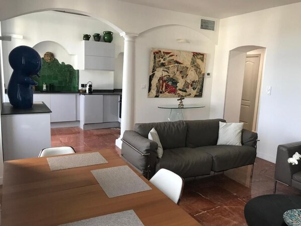 A modern living room with a sofa, a coffee table, and a dining table. There is a kitchen in the background with white cabinets and a green backsplash.
