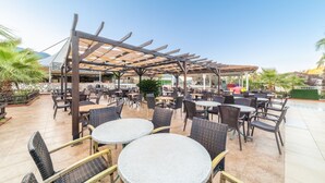 An outdoor patio area with round tables and chairs, a pergola structure, and a pool in the background. - Sahra Su Holiday Village & SPA (Fethiye)