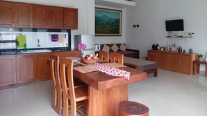 A modern kitchen with wooden cabinets, a dining area with a wooden table and chairs, a sofa, and a painting on the wall. - Just outside Ubud, peaceful 3BR Villa with rice field views. (Tampaksiring, Gianyar Regency)