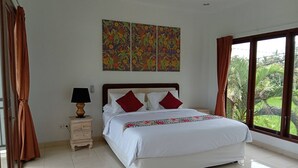 A bedroom with a large bed, a nightstand with a lamp, a window with curtains, and a wall with three paintings. - Just outside Ubud, peaceful 3BR Villa with rice field views. (Tampaksiring, Gianyar Regency)