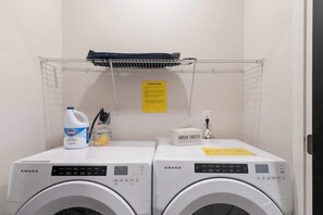 Two AMANA washing machines in a laundry room with a shelf above holding laundry baskets.