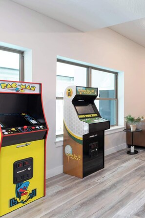 Two arcade machines, one labeled 'PAC-MAN' and the other 'Golden Tee 3D-GOLF,' in a room with wooden flooring and large windows.