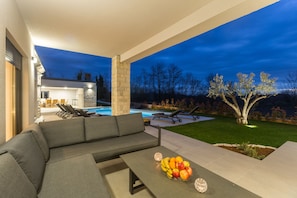 Et udendørs opholdsområde med en moderne sofa, et spisebord med frugt og en pool i baggrunden.