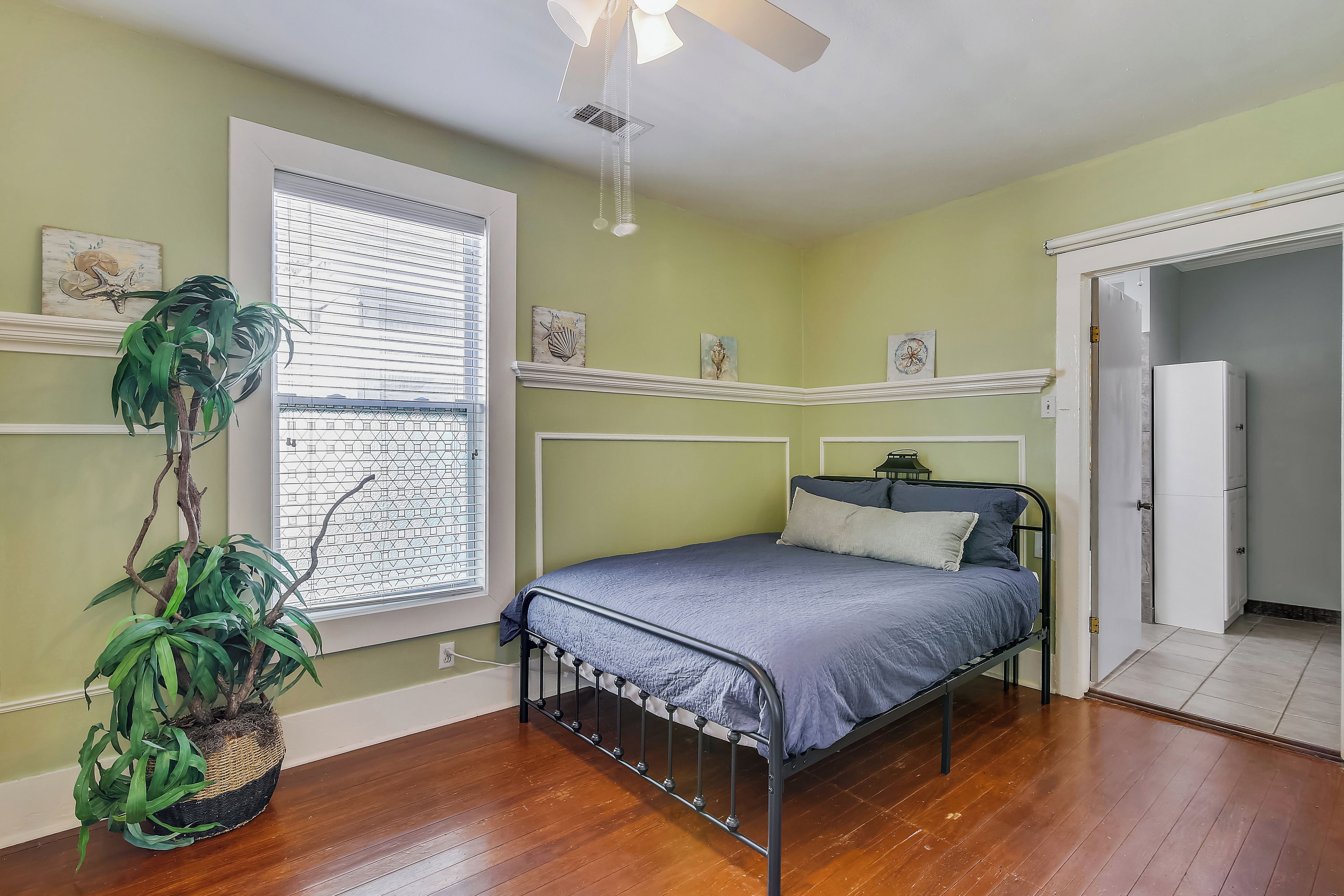 A bedroom with a bed, a potted plant, a window with blinds, and a door leading to another room.