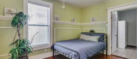 A bedroom with a bed, a potted plant, a window with blinds, and a door leading to another room.