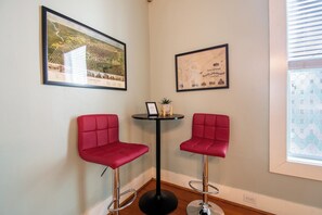 A corner room with two red bar stools, a small table with a plant, and two framed pictures on the wall.