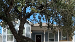 A two-story house with a garage, a tree in front, and a fence. - A Bended Oak Bungalow by Ryson Vacation Rentals (Galveston)