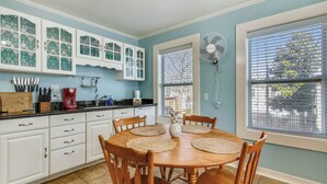 A kitchen with white cabinets, a dining table with placemats, and a fan near the window. - A Bended Oak Bungalow by Ryson Vacation Rentals (Galveston)