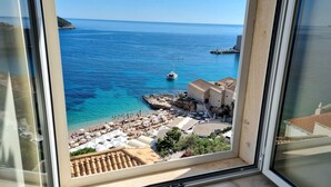 Une vue sur la côte, depuis une fenêtre donnant sur une plage avec des parasols, un bateau et des bâtiments historiques.